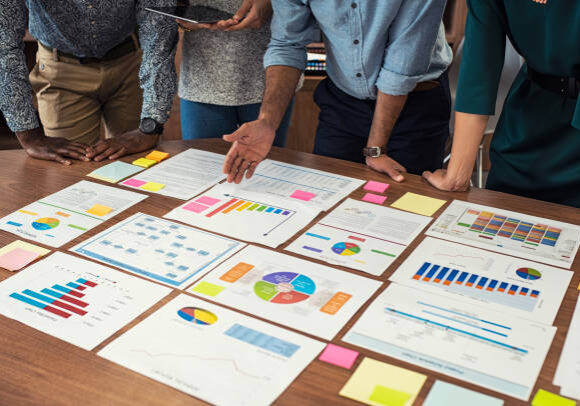 Financial and business documents on table with multiethnic hands working on it. Latin business manager with colleagues working on new startup project. Closeup business man and businesswoman hands understanding pie and bar graphs during meeting.